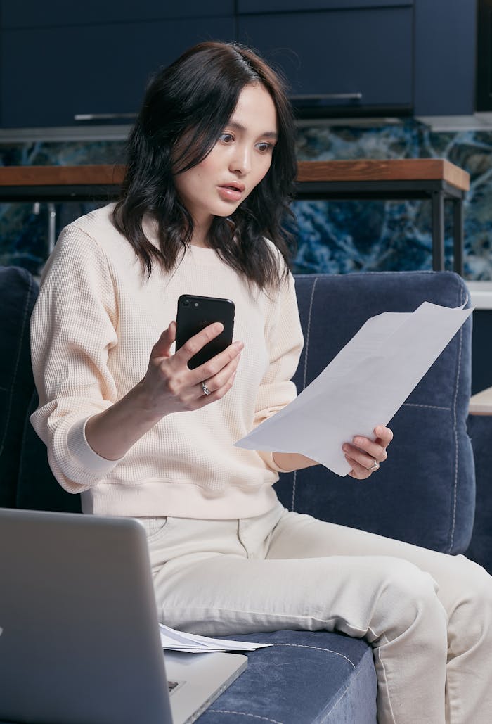 services-01 A young woman uses her smartphone and laptop to manage finances, appearing focused and concerned.