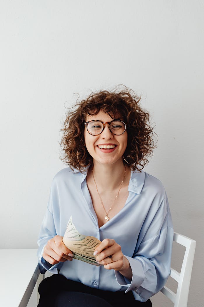 services-03 Cheerful woman with curly hair and glasses holding US dollars, sitting indoors.